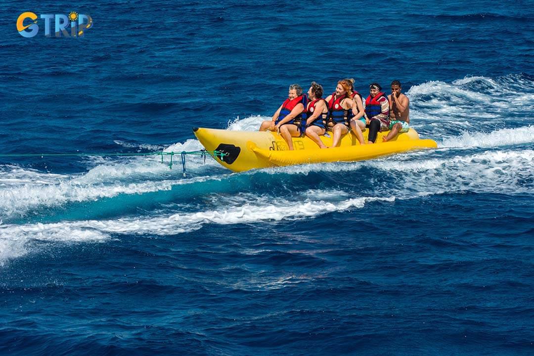 Tourists have fun and enjoy riding on a red and white inflatable banana that carries a scooter across the sea